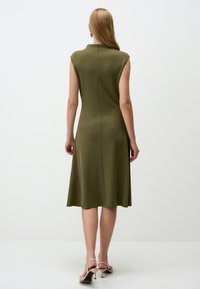 Olivegrünes ärmelloses Kleid mit einem taillierten Oberteil und einem ausgestellten Saum, aus glattem Stoff gefertigt. Dezentes Nahtdetail im Rücken. Sichtbare beige Riemchenheels.