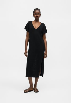 Kobieta stojąca, ubrana w czarną midi sukienkę z krótkim rękawem, rozcięciem po boku i czarne klapki, na jednolitym białym tle.