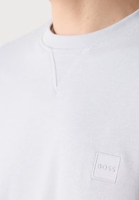 Gros plan d’un t-shirt blanc à col rond avec un petit logo carré embossé indiquant "BOSS" sur la poitrine.