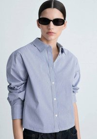 Blue and white striped button-up shirt with long sleeves, featuring subtle textured dots and a classic collar, paired with black sunglasses.
