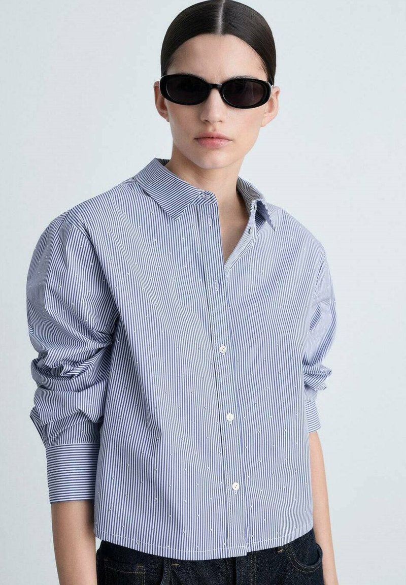 Blue and white striped button-up shirt with long sleeves, featuring subtle textured dots and a classic collar, paired with black sunglasses.