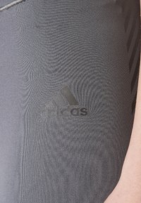 Šedé sportovní legíny vyrobené z hladké, pružné látky. Černé logo Adidas umístěné na horní části stehna, s jemnými texturními detaily.
