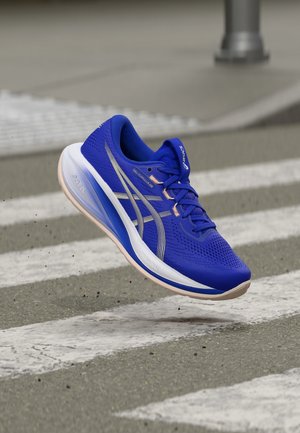 Scarpa da corsa blu con suola bianca e beige sospesa in aria sopra un attraversamento pedonale su una strada in asfalto grigio.