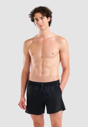 Joven con cabello rizado que lleva pantalones cortos de baño negros, de pie con una mano en el bolsillo contra un fondo gris liso.