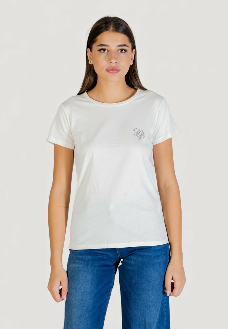 Biały bawełniany t-shirt z krótkimi rękawami, okrągłym dekoltem i srebrnym logo na lewej piersi. W połączeniu z niebieskimi jeansami.