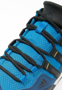 Blå sportig sko med texturerad mesh-ovandel, svarta skosnören och svart hälkappa. Märkning synlig på sidan och hälen.