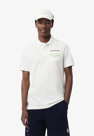 Polo bianco con superficie testurizzata, dotato di logo ricamato verde, strisce di accento rosse e blu navy, abbinato a un cappellino bianco.