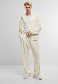 Traje deportivo color crema hecho de una tela suave, que cuenta con una chaqueta con cremallera y bolsillos, y pantalones de cintura elástica a juego. Detalle sutil del logo.