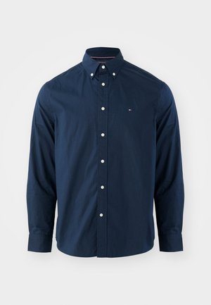 Chemise à manches longues bleu marine avec un col boutonné, boutons blancs et un petit logo sur la poitrine. Texture de tissu lisse.