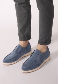 Niebieskie zamszowe buty typu slip-on z zaokrąglonym nosem, gumową podeszwą i przeszyciami. Noszone z szarymi spodniami z podwiniętymi nogawkami.