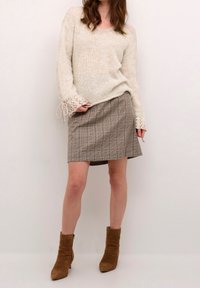 Beżowy sweter z dzianiny z rękawami wiązany na sznurki, w połączeniu z brązową spódnicą w kratę. Modelka nosi jasnobrązowe botki za kostkę.