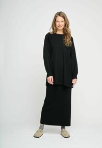 Svart långärmad topp med en lös passform, ihopparad med en matchande svart kjol i ankel-längd. Modellen bär lätta tofflor och grå strumpor.