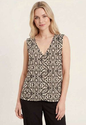 Femme aux cheveux blonds portant un haut beige sans manches avec des motifs géométriques noirs et un pantalon noir, debout devant un fond uni.