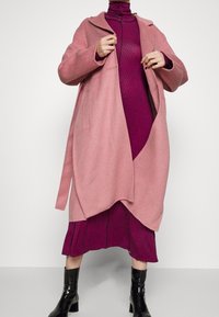 Pink wool coat with an open front, featuring a collar and side pockets, layered over a textured magenta dress, paired with black ankle boots.