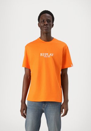 Camiseta de algodón naranja con un logo blanco "REPLAY SINCE 1981" en el pecho, escote redondo y mangas cortas, combinada con jeans azules.