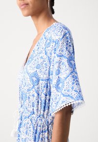 Robe à imprimé floral avec des couleurs bleues et blanches, manches courtes évasées, taille cintrée avec cordon de serrage, et délicate finition en dentelle.