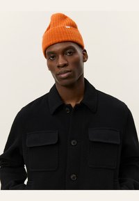 Abrigo negro texturizado con dos bolsillos de parche y cierre de botones, combinado con un gorro acanalado naranja brillante. Fondo neutro.