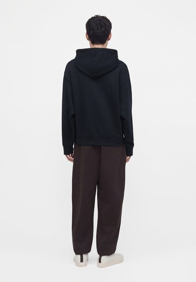 Personne debout, de dos, portant un sweat à capuche noir, un pantalon large foncé et des baskets blanches sur un fond blanc uni.