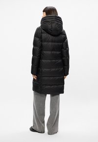 Cappotto imbottito nero con cappuccio removibile, caratterizzato da un design trapuntato e maniche lunghe. Abbinato a pantaloni grigi a gamba larga.