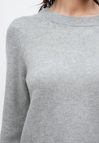 Pull en tricot gris clair doux avec un col rond porté par une personne avec une zone visible du cou et des épaules.
