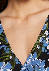 Gros plan sur le haut de la poitrine et le cou d'une personne portant un vêtement noir avec des motifs floraux bleus et blancs et des feuilles vertes.