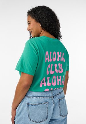 Kvinde med krøllet hår iført en grøn skjorte med teksten "ALOHA CLUB" i pink bogstaver, kombineret med lyseblå denimjeans.