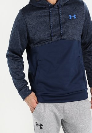 Mężczyzna w granatowej i ciemnoniebieskiej bluzie z kapturem ze sznurkami oraz szarych spodniach dresowych, oba z logo Under Armour. Ręce w kieszeniach bluzy i spodni.