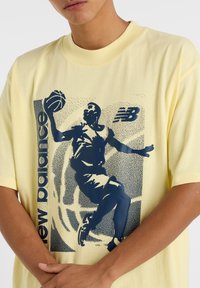 T-shirt en coton jaune avec un imprimé graphique bleu représentant un joueur de basket-ball et le texte "new balance" dans une police audacieuse et moderne.