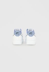 Fehér Nike Air sportcipő világos lila sarokdíszítéssel és fekete logóval. Sima bőr textúra, lekerekített orr, mintás gumi talp.