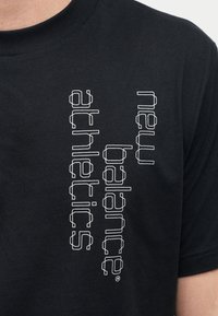 Μαύρο μπλουζάκι με ασπρόμαυρο περίγραμμα κειμένου «nature balance athletics» τυπωμένο κάθετα στο στήθος, που φοριέται από άτομο με ανοιχτό χρώμα δέρματος.