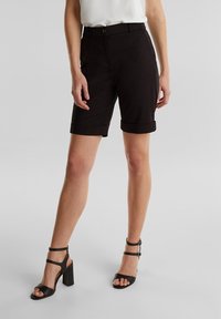 Nainen, joka pitää yllään mustia vyötärölle käännettyjä polvipituisia shortseja ja mustia korkeakorkoisia sandaaleja, seisoo tasaisella vaalealla taustalla.