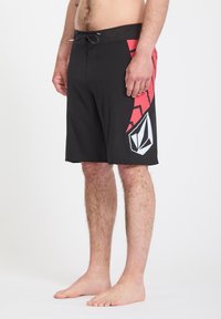 Homme portant un short de bain noir avec un motif géométrique rouge et un logo blanc sur la jambe droite, debout pieds nus devant un fond blanc.