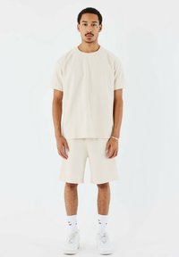 Camiseta de manga corta beige con cuello redondo combinada con pantalones cortos a juego. La tela tiene un aspecto texturizado. Zapatillas blancas y calcetines cortos completan el atuendo.