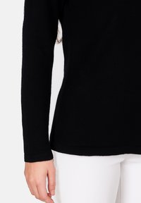 Schwarzes Langarmshirt aus einem weichen, strukturierten Stoff, mit figurnaher Passform, abgerundetem Saum und engen Bündchen, kombiniert mit weißen Hosen.