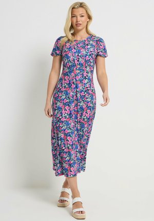 Femme debout portant une robe midi bleue à fleurs roses et des sandales compensées blanches sur un fond clair uni.