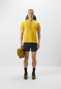 Gelbes kurzärmliges Hemd mit Reißverschlusskragen, schwarze Shorts, trägt eine braune Tasche. Schwarze Socken und Schuhe mit Tarnmuster runden das Outfit ab.