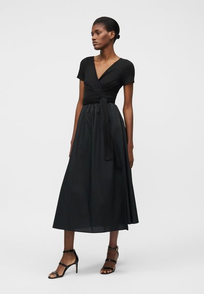 Γυναίκα που στέκεται φορώντας μαύρο τοπ τύπου φάκελος με V-λαιμόκοψη και κοντά μανίκια, μαζί με μαύρη, αέρινη midi φούστα με ψηλή μέση και μαύρα σανδάλια με τακούνι.