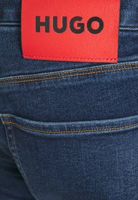 Jeans de mezclilla azul oscuro con costuras doradas, una etiqueta de cuero roja con "HUGO" estampado en negro, destacando la identidad de la marca.