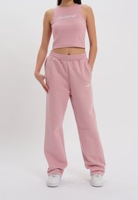 Donna che indossa un top corto senza maniche rosa e pantaloni sportivi larghi abbinati, con scarpe da ginnastica bianche, in piedi con le mani nelle tasche.