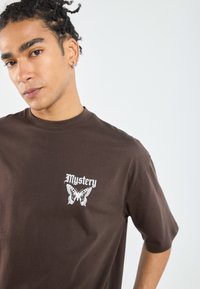 Hnědá bavlněná tričko s bílým grafickým potiskem slova "Mystery" a motýlem na levé straně hrudi. Krátké rukávy, design s kulatým výstřihem.