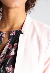 Rosa texturerad blazer med krage, kombinerad med en blus med blommönster i mörka färger som har vita och röda blommor.