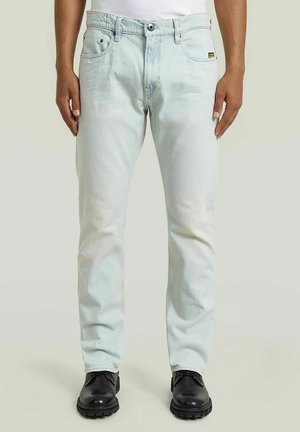 Homme portant un jean bleu clair à coupe droite avec poches avant et chaussures noires à lacets sur fond uni.