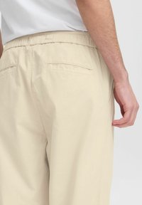 Beige elastic midjebukser med to baklommer og en avslappet passform. Stoffet virker lett og glatt i tekstur.