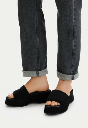 Nohy v černých pantoflích s platformou, doplněné o svinuté tmavě šedé džíny na bílém pozadí.