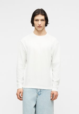 Giovane con capelli castani di media lunghezza che indossa una maglia bianca a maniche lunghe e jeans azzurri, in piedi davanti a uno sfondo neutro.