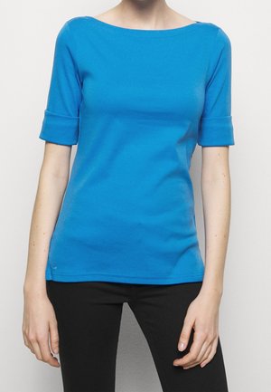 Personne portant une chemise à manches courtes bleu vif avec des poignets retroussés et un pantalon noir, debout devant un fond clair uni.