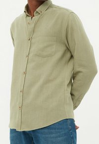 Camisa de botonadura en verde claro, hecha de tela texturizada, con un bolsillo en el pecho, pequeño cuello y botones de madera, combinada con jeans azules.