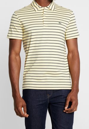Homme portant un polo jaune pâle à manches courtes avec de fines rayures horizontales noires et un jean bleu foncé, debout les bras détendus.