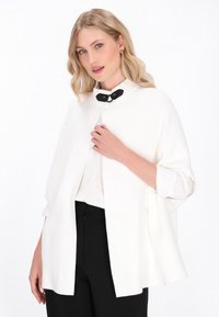 Fehér köpeny fekete gombos záródással, magas gallérral és laza ujjakkal, fehér gombos ingre és fekete nadrágra rétegezve.