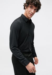 Zwarte, lange mouwen, blouse met knopen, een brede kraag, een gladde textuur en een aansluitend ontwerp. Draagt zwarte broek en bijpassende riem.